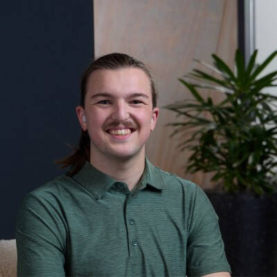An image of Zach Williams, a Blue Nucleus Developer. Zach is sitting by a small plant behind him, and wearing a green button-up shirt.